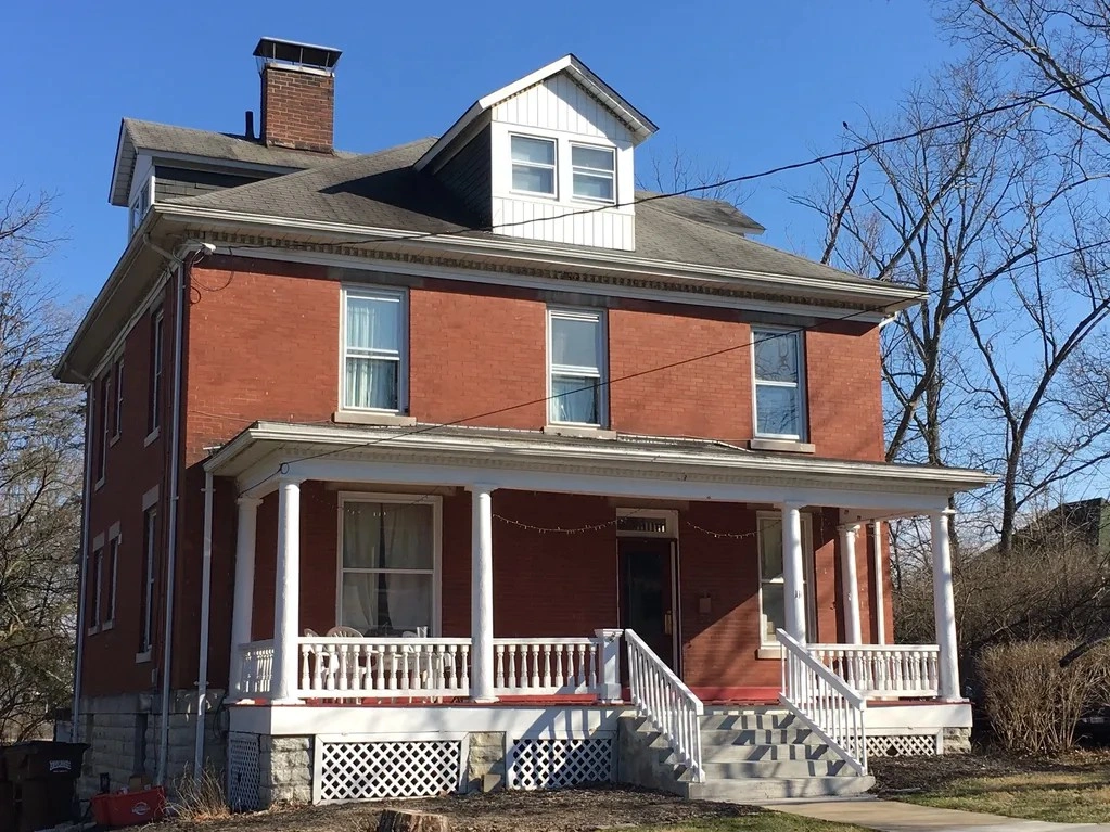 Historic red-brick Victorian house with white porch columns, dormer windows, chimney, front steps, and leafless trees under a blue sky.